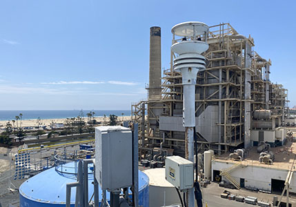 A Magellan MX500 Weather Station is shown in the foreground, with the power generation plant in the background near a beach and a blue sky.