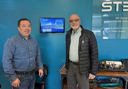 Nader and Fabio stand in the lobby of Stevens Water Monitoring Systems against a blue wall and a screen welcoming Nader and Martha.